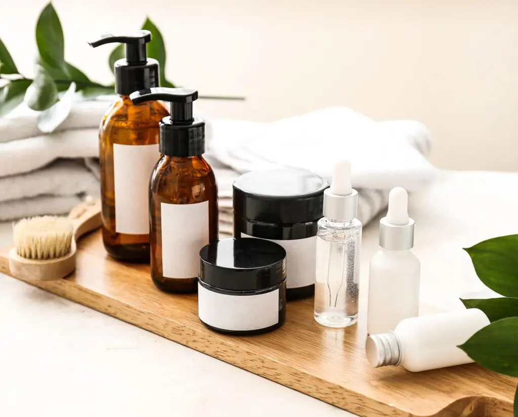 A collection of beauty and personal care products displayed on a wooden tray, including amber pump bottles, cream jars, serum droppers, and skincare containers, styled with soft towels and greenery