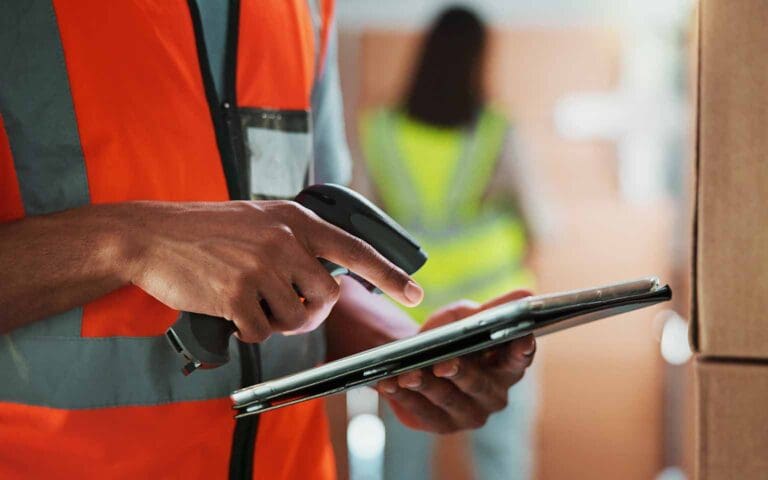 Warehouse worker scans packages with a barcode scanner and tablet, illustrating how Amazon sellers are managing inventory and navigating supply chain challenges intensified by tariffs