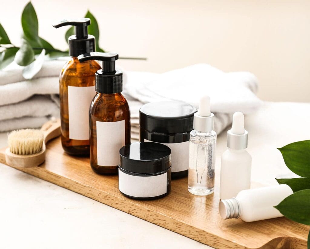 A collection of beauty and personal care products displayed on a wooden tray, including amber pump bottles, cream jars, serum droppers, and skincare containers, styled with soft towels and greenery